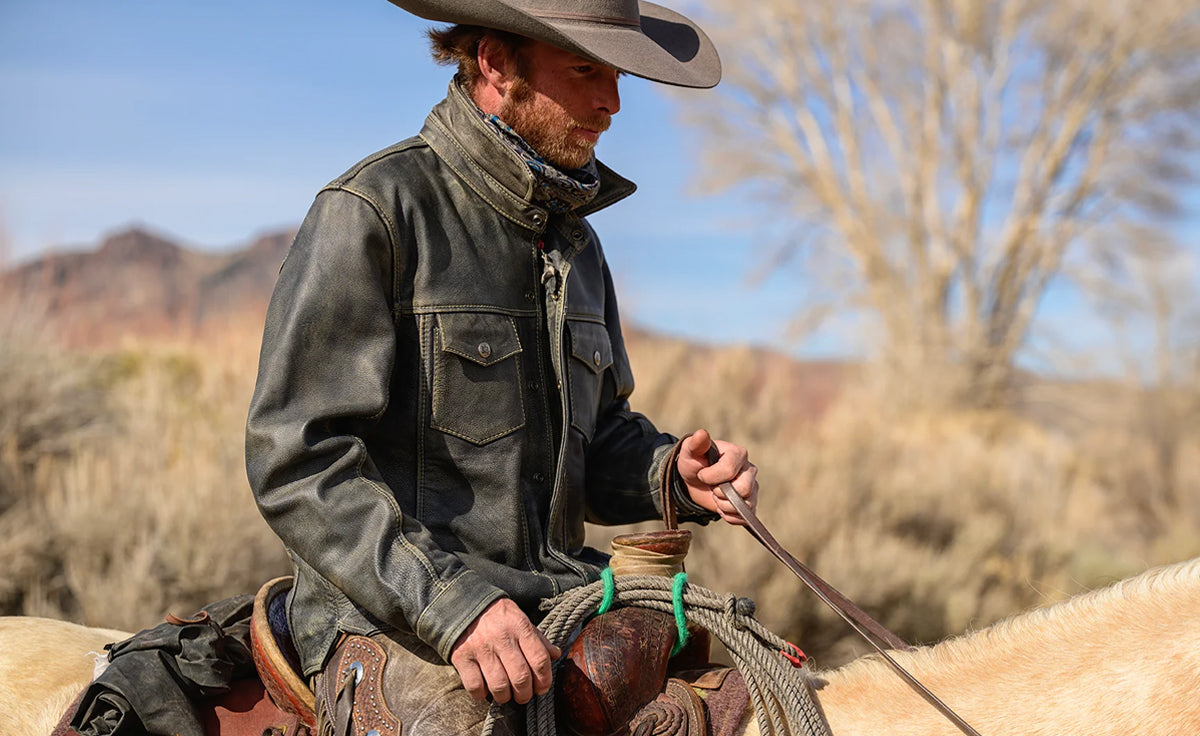Man wearing a distressed black cowboy leather jacket while riding a horse on a ranch