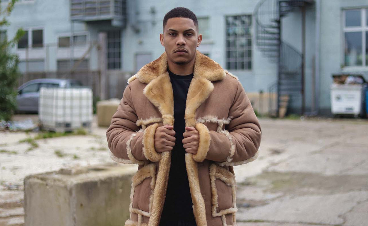 Man wearing a tan shearling coat with a wide wool collar and exposed shearling trim outdoors.