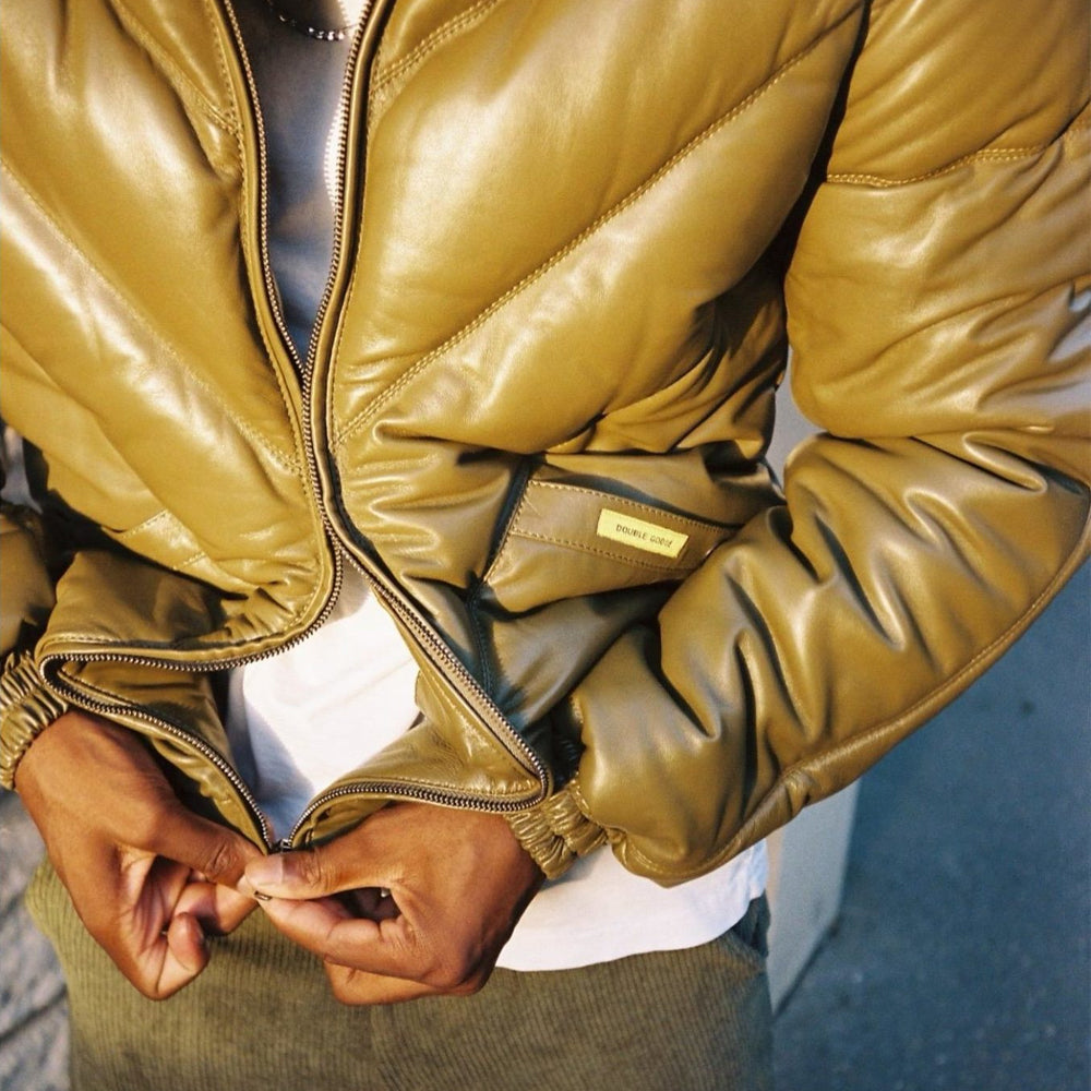 Men’s olive leather V-bomber worn open, highlighting padded chevrons, pocket label, zipper and elasticized cuffs