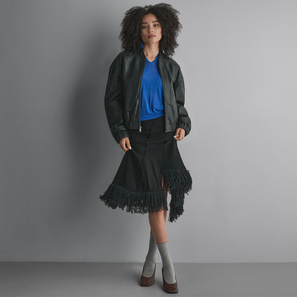 Full-length styling view of a relaxed black leather bomber jacket worn with a blue knit top and fringed midi skirt.